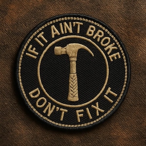 May include: Circular embroidered patch with the phrase "IF IT AIN'T BROKE DON'T FIX IT" in gold thread on a black background. A gold hammer is in the centre. The patch is set against a brown fabric backdrop.