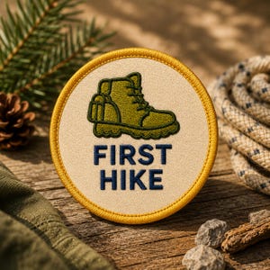 May include: A circular embroidered patch with a cream-colored background and a yellow stitched border. The patch features an olive green hiking boot graphic above the dark blue text FIRST HIKE, centered in a bold, sans-serif typeface.