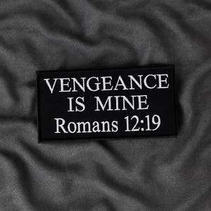 Op de afbeelding: Een zwart rechthoekig embleem met wit geborduurde tekst. De tekst luidt "VENGEANCE IS MINE Romans 12:19". De patch is geplaatst tegen een grijze, getextureerde achtergrond.