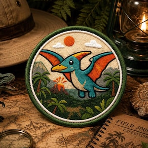Puede incluir: Un parche bordado circular que presenta un pterodáctilo de dibujos animados con piel azul y alas naranjas volando sobre un paisaje prehistórico. La escena incluye palmeras verdes, un volcán humeante, un sol naranja brillante y nubes blancas.