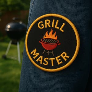 May include: A round black patch with the words "GRILL MASTER" in yellow, surrounding a red grill with orange flames. The patch is on a blue denim background, with a blurred grill in the background.