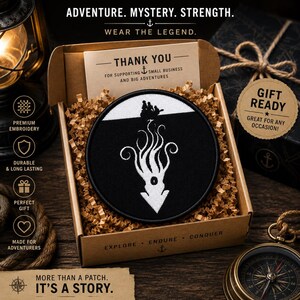 May include: A circular embroidered patch featuring a white silhouette of a ship on the surface above a large white kraken or squid creature against a black background, presented inside a brown cardboard gift box filled with paper filler.