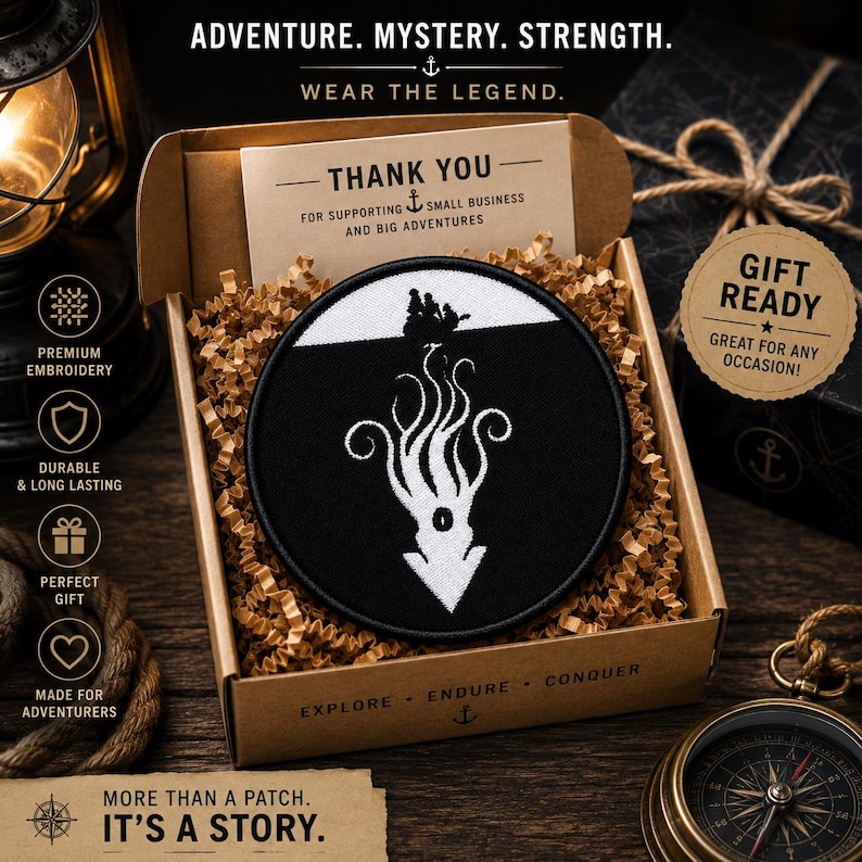 May include: A circular embroidered patch featuring a white silhouette of a ship on the surface above a large white kraken or squid creature against a black background, presented inside a brown cardboard gift box filled with paper filler.