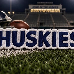 May include: A rectangular embroidered patch featuring the word HUSKIES in bold dark blue capital letters with a thick white border. The patch has a textured fabric surface and is positioned horizontally with a clean, stitched edge finish.