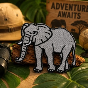 May include: A gray embroidered elephant patch with black outlines. The patch is on a piece of wood, with a safari hat, binoculars, and a globe in the background. The sign in the background reads "ADVENTURE AWAITS".