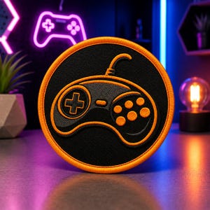 Könnte beinhalten: Runder schwarzer Aufnäher mit einem gestickten orangefarbenen Controller-Design. Der Controller hat ein Kreuz, Knöpfe und ein Kabel. Der Aufnäher hat einen orangefarbenen Rand. Der Hintergrund umfasst ein Neon-Controller-Schild und eine kleine Pflanze.