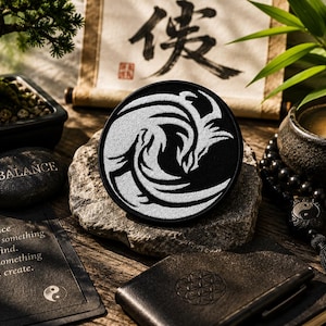 Pode incluir: Um emblema circular preto com um design bordado em branco de duas criaturas estilizadas entrelaçadas que lembram dragões. O emblema está centrado sobre uma pedra texturizada, rodeado por um bonsai, uma vela acesa, um caderno preto e um cartão com o texto "Balance is not something you find. It's something you create."