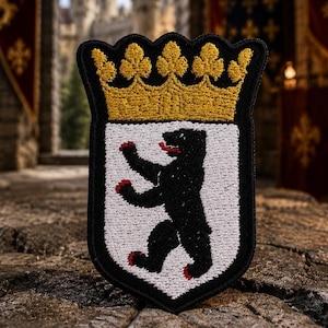 Könnte beinhalten: Ein schildförmiger, bestickter Aufnäher mit einem schwarzen Bären, der auf seinen Hinterbeinen steht, mit roten Krallen und roter Zunge auf weißem Grund, gekrönt von einer goldgelben Krone, alles mit einem dicken schwarzen Rand.