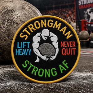 Op de afbeelding: Een ronde geborduurde patch met een zwarte achtergrond en een gele rand. Het toont een wit silhouet van een persoon die een steen optilt, met de tekst "STRONGMAN" in het geel, "LIFT HEAVY" in het blauw, "NEVER QUIT" in het oranje en "STRONG AF" in het groen.