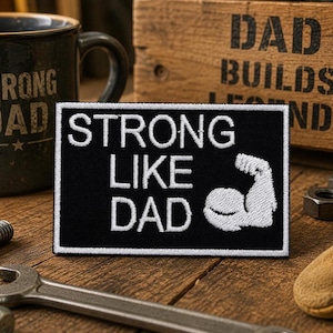 May include: A rectangular black embroidered patch featuring white text that reads STRONG LIKE DAD and a white graphic of a flexed bicep muscle. The patch has a clean white border and is designed for attachment to fabric items or clothing.