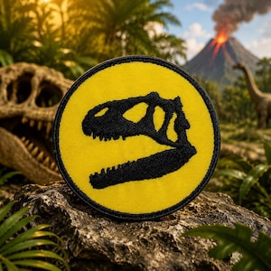 May include: Round yellow patch with a black dinosaur skull design. The patch has a black border and is set against a background of a prehistoric landscape with a volcano and dinosaurs. The patch is likely an iron-on or sew-on patch.