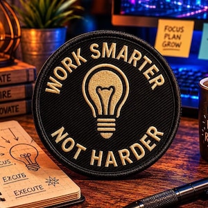 May include: A black circular patch with the text "WORK SMARTER NOT HARDER" in gold lettering, surrounding a gold lightbulb graphic. The patch is on a wooden surface, with a notebook, pen, and coffee mug nearby. The notebook has a lightbulb diagram.