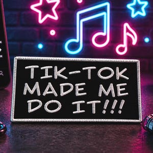 May include: A rectangular black embroidered patch featuring the white stitched text TIK-TOK MADE ME DO IT!!! with a white border. The patch is positioned on a dark surface alongside a smartphone, a small potted succulent, and a spiral notebook.