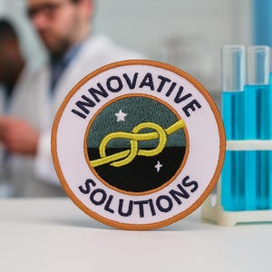 May include: A circular embroidered patch with a white border and an orange edge. The center features a yellow knot on a split green and black background with two small white stars. The text "INNOVATIVE SOLUTIONS" is stitched in dark blue.
