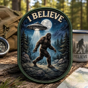 May include: An oval patch with a Bigfoot illustration under a UFO and the text "I BELIEVE". The background depicts a forest scene with a full moon. The patch is surrounded by a green border.