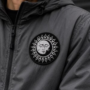 May include: A charcoal grey hooded jacket featuring a circular embroidered patch on the chest. The patch displays a white sun and moon face design with flame-like rays against a black background, complete with a front zip and drawstring hood.
