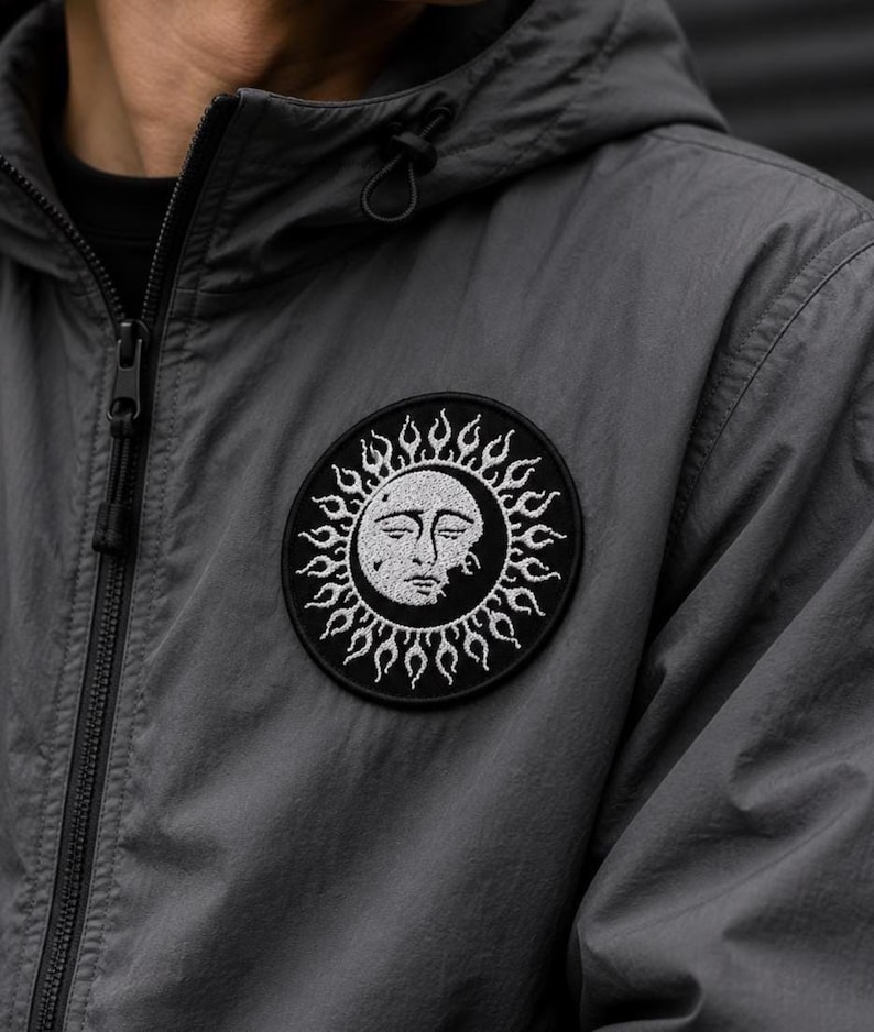 May include: A charcoal grey hooded jacket featuring a circular embroidered patch on the chest. The patch displays a white sun and moon face design with flame-like rays against a black background, complete with a front zip and drawstring hood.