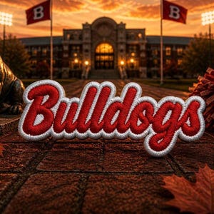 May include: A red and white embroidered patch featuring the word Bulldogs in a cursive script. The patch is positioned on a brick surface with fallen autumn leaves, a bronze bulldog statue, and a red pom-pom nearby, with a campus building in the background.