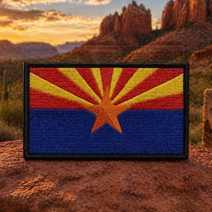 May include: A rectangular embroidered patch featuring the Arizona state flag design. The top half displays thirteen alternating red and yellow rays, while the bottom half is solid blue. A large copper-colored five-pointed star is centered.