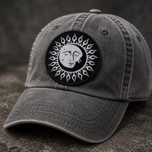 May include: A charcoal grey baseball cap with a distressed finish featuring a circular black patch on the front. The patch displays an embroidered white sun design with a human face and radiating flame-like rays surrounding the central sun icon.
