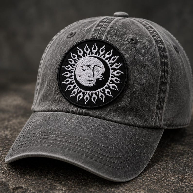 May include: A charcoal grey baseball cap with a distressed finish featuring a circular black patch on the front. The patch displays an embroidered white sun design with a human face and radiating flame-like rays surrounding the central sun icon.