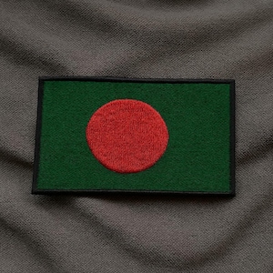 Könnte beinhalten: Ein rechteckiger Aufnäher mit der Flagge von Bangladesch. Die Flagge hat einen dunkelgrünen Hintergrund mit einem großen, zentrierten roten Kreis. Der Aufnäher ist von einem schwarzen Rand umgeben.