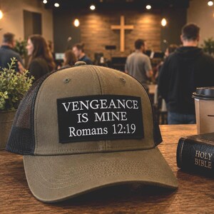 Op de afbeelding: Olijfgroene truckerpet met een zwarte mesh achterkant en een rechthoekige patch met de tekst "VENGEANCE IS MINE Romeinen 12:19" in het wit. Een koffiekopje met een kruis en een gesloten Bijbel liggen ook op de tafel.