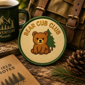 May include: A circular embroidered patch with a tan background and a dark green border. The patch features a brown bear cub illustration, a green pine tree, and the text BEAR CUB CLUB embroidered in gold thread along the top edge.