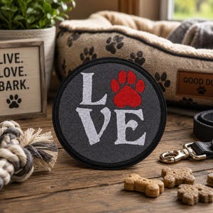May include: A circular dark gray embroidered patch featuring the white text LOVE with a red paw print replacing the letter O. The patch has a black stitched border and is displayed on a wooden surface alongside dog-themed items and treats.