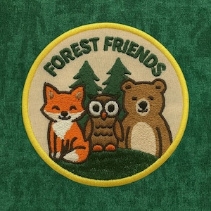 Pode incluir: Patch bordado com uma raposa, uma coruja e um urso, com o texto "FOREST FRIENDS" num design circular. O patch tem um fundo bege com árvores verdes e uma borda verde, sobre um tecido verde.