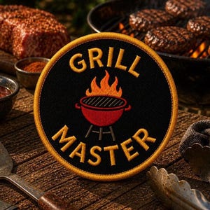 May include: A circular embroidered patch with a black background and a gold border. The patch features an orange and red graphic of a grill with flames and the yellow embroidered text GRILL MASTER arranged in an arc above and below the grill.