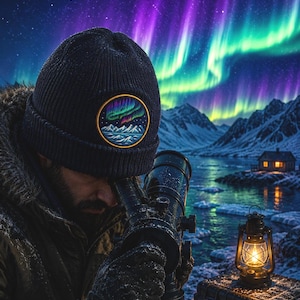 May include: A dark navy blue knit beanie featuring a circular embroidered patch that depicts a vibrant aurora borealis over snow-capped mountains. The beanie is worn by an individual looking through a black telescope in a snowy landscape.