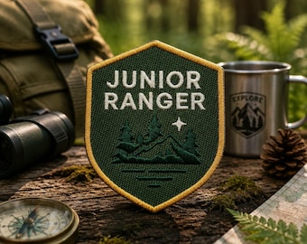 May include: A green and gold Junior Ranger patch with a mountain and tree design. The patch is on a wooden surface with a compass, metal mug, and map. The mug has the word "EXPLORE" and a mountain graphic.
