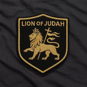 Könnte beinhalten: Gestickter Aufnäher mit einem goldenen Löwen, Krone und Kreuz auf einem schwarzen Schild. Der Text "LION OF JUDAH" ist in Gold über dem Löwen. Der Aufnäher hat einen goldenen Rand und ist auf dunklem Hintergrund platziert.
