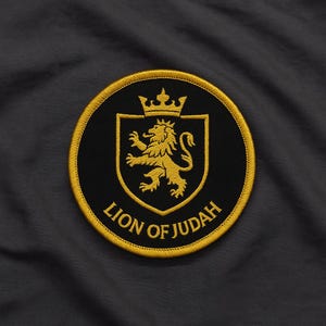 Peut inclure: Écusson brodé circulaire noir avec un lion doré couronné. Le texte "LION OF JUDAH" est inscrit en arc de cercle. Le patch est bordé d'or et posé sur un tissu gris foncé.