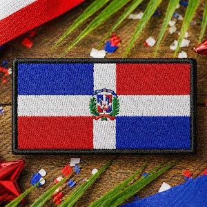 Może przedstawiać: Haftowana naszywka z flagą Dominikany, z białym krzyżem dzielącym czerwone i niebieskie kwadraty. Prostokątna naszywka ma czarną obwódkę, otoczoną czerwonymi gwiazdkami i wstążkami.