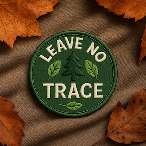 May include: A round, green embroidered patch with the words "LEAVE NO TRACE" in white. The patch features a green pine tree and three green leaves. The patch is surrounded by a green border and set against a brown background with autumn leaves.