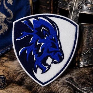 May include: A shield-shaped embroidered patch featuring a stylized blue lion head design on a split white and dark blue background. The patch has a thick white border and is constructed with dense thread work, showcasing a sharp, angular aesthetic.