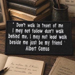 May include: A rectangular black embroidered patch featuring white text that reads: Don't walk in front of me I may not follow don't walk behind me. I may not lead walk beside me just be my friend Albert Camus. The patch has a stitched border.