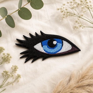 May include: An embroidered patch shaped like an eye with long black eyelashes. The iris is a vibrant blue with a black pupil and a white highlight. The sclera is white, and the surrounding area is black, creating a sharp, graphic contrast.