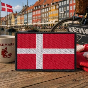 Puede incluir: Un parche rojo y blanco con la bandera danesa, una cruz blanca sobre fondo rojo. A la izquierda, una taza con la palabra "DANMARK" y una silueta roja. El fondo muestra una bicicleta y edificios.