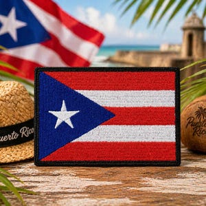 Può includere: Una toppa rettangolare con la bandiera di Porto Rico, con un triangolo blu, una stella bianca e strisce rosse e bianche. Sono visibili anche un cappello di paglia con "Puerto Rico" su una fascia nera e una noce di cocco con "Puerto Rico".
