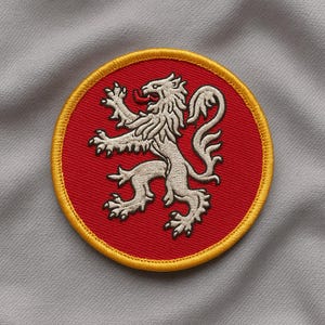 Puede incluir: Parche bordado circular con un león blanco rampante sobre fondo rojo. El león presenta detalles finos y está rodeado por un borde amarillo. Ideal para ropa o accesorios.