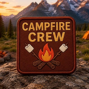 Puede incluir: Un parche cuadrado bordado de color marrón con el texto amarillo CAMPFIRE CREW sobre un gráfico de una llama central naranja y amarilla, leños marrones y dos malvaviscos blancos en palos inclinados a ambos lados del fuego.