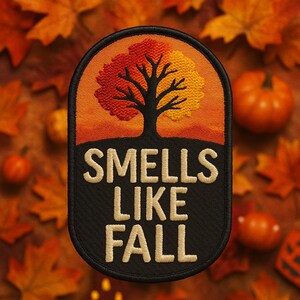 Puede incluir: Parche bordado con temática otoñal. El parche presenta un árbol con hojas rojas y amarillas sobre un cielo naranja. El texto "Smells Like Fall" está bordado en beige sobre un fondo negro. Calabazas y hojas de otoño rodean el parche.