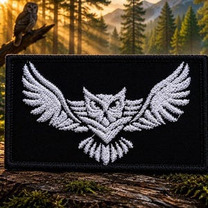 May include: A rectangular black patch featuring a white embroidered design of an owl with its wings spread wide. The owl is depicted in a stylised, symmetrical manner with detailed feather patterns on the wings and a sharp, focused gaze.
