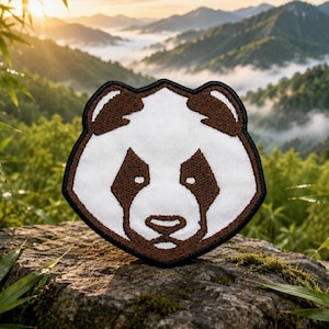 May include: A shield-shaped embroidered patch featuring the stylized face of a panda. The design uses brown thread for the ears, eye patches, nose, and outer border, contrasted against a white thread fill for the face and forehead details.