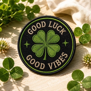 May include: A circular embroidered patch with a black background and a green four-leaf clover at the center. The text GOOD LUCK is stitched in beige above the clover, and GOOD VIBES is stitched in beige below, with small green star accents.
