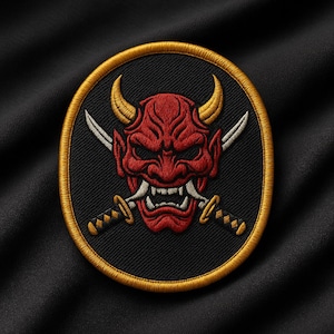 May include: Embroidered patch featuring a red Oni mask with yellow horns and two crossed swords. The mask is set against a black background within a gold oval border. The patch is likely for jackets or bags.