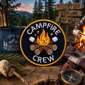 May include: A round, black patch with the words "Campfire Crew" in white, featuring a campfire design with marshmallows. A blue enamel mug with the text "Life is Better Outdoors" is also visible. The scene evokes camping and outdoor adventure.